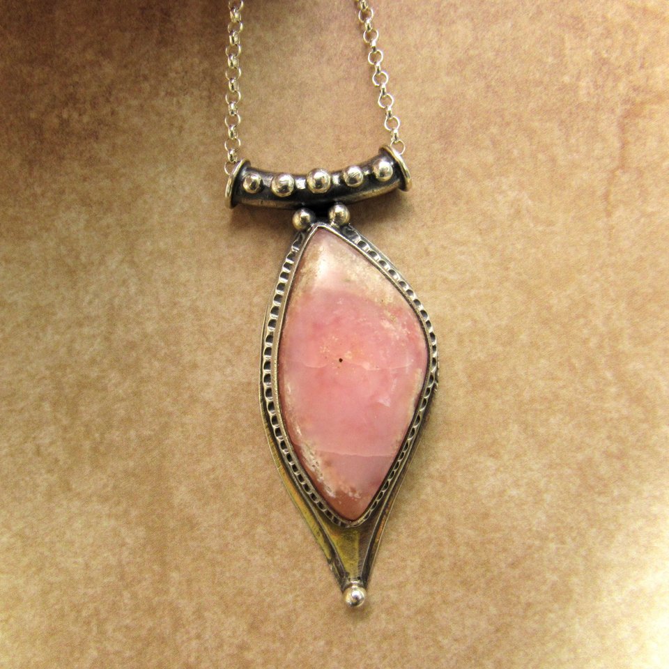 Soft elegance meets artisan craftsmanship in this one of a kind handcrafted pendant featuring a beautifully pastel pink opal set in textured Argentium® sterling silver. The unique, petal-shaped stone in a petal shaped setting radiates gentle warmth in a soothing blush hue—perfect for those who adore subtle statement pieces with soulful charm. 2.25" long x 7/8" wide.