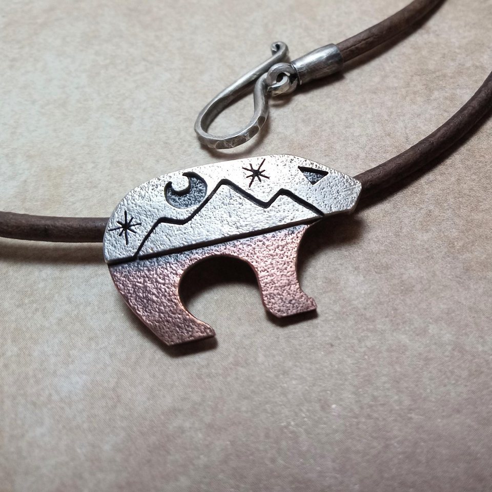 Artisan made Copper and silver bear pendant on leather cord with SS handmade findings. 1.25" long and .75" tall. Shown against light brown background.