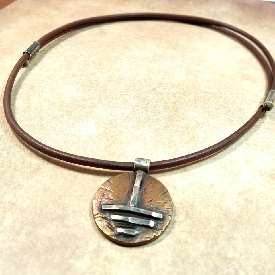 Full view of unisex bronze and silver grounding symbol pendant with adjustable leather cord, photo in hand.