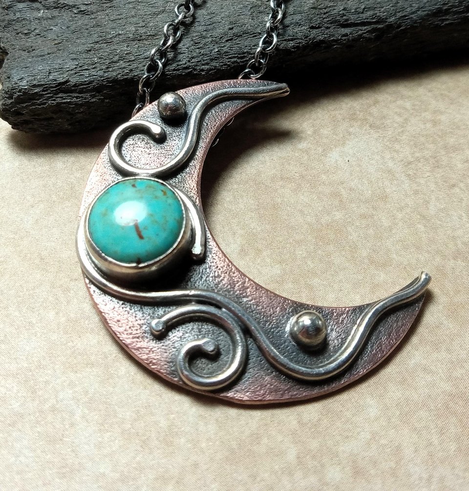Close up of copper and silver moon pendant with turquoise cabochon, photo against rock on beige background.