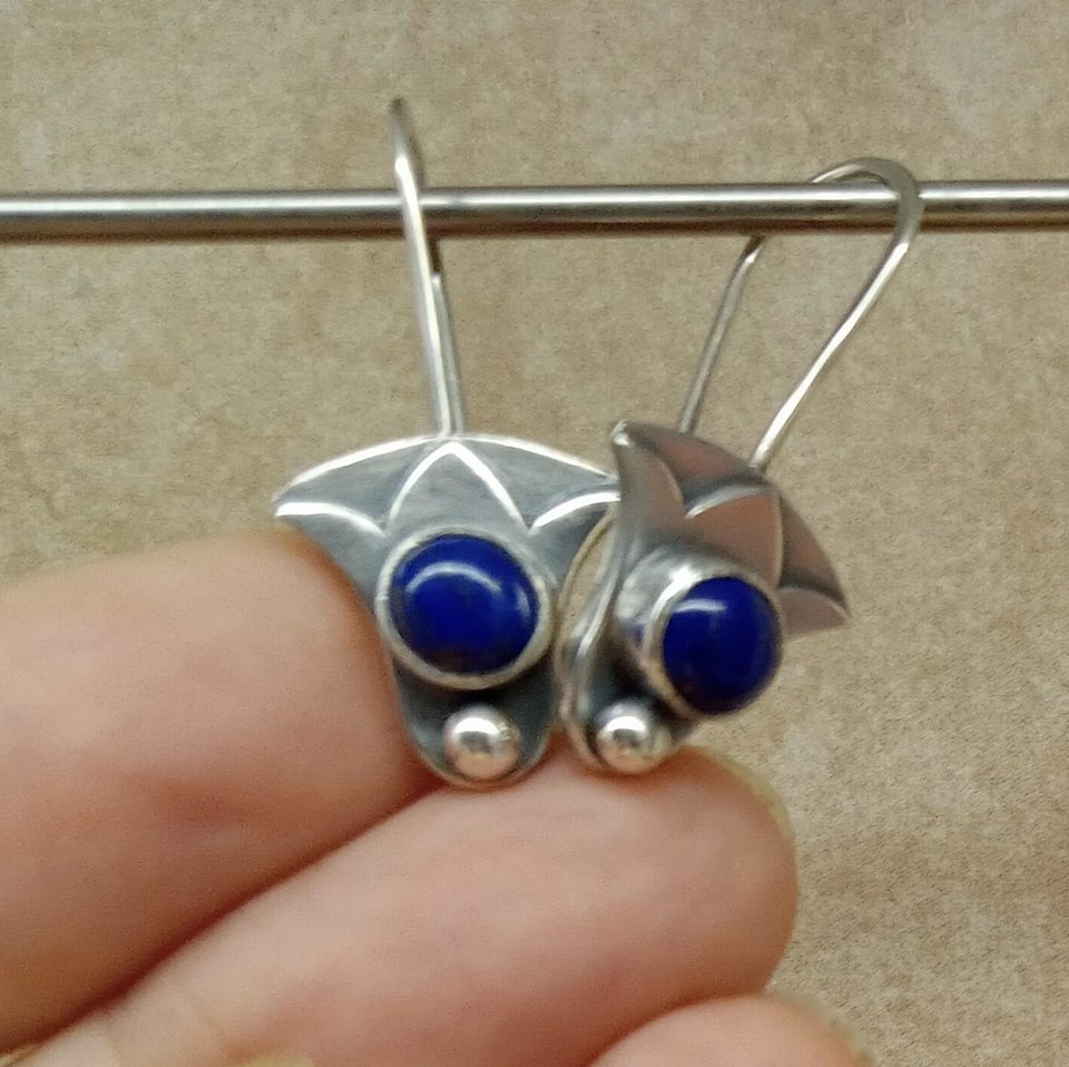 Pair of lotus earrings with lapis lazuli displayed on fingers against neutral surface.