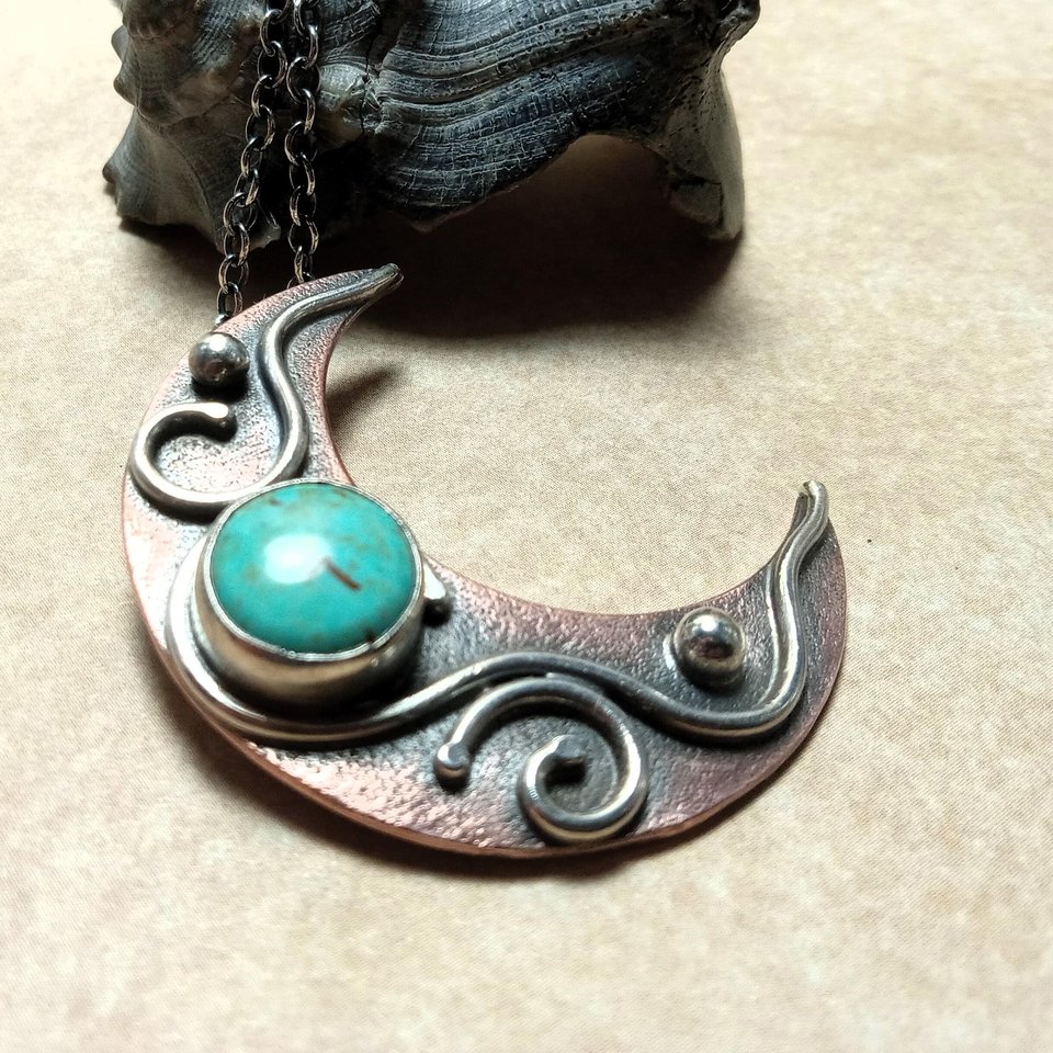 Copper and silver moon pendant set with turquoise against black shell on beige background.