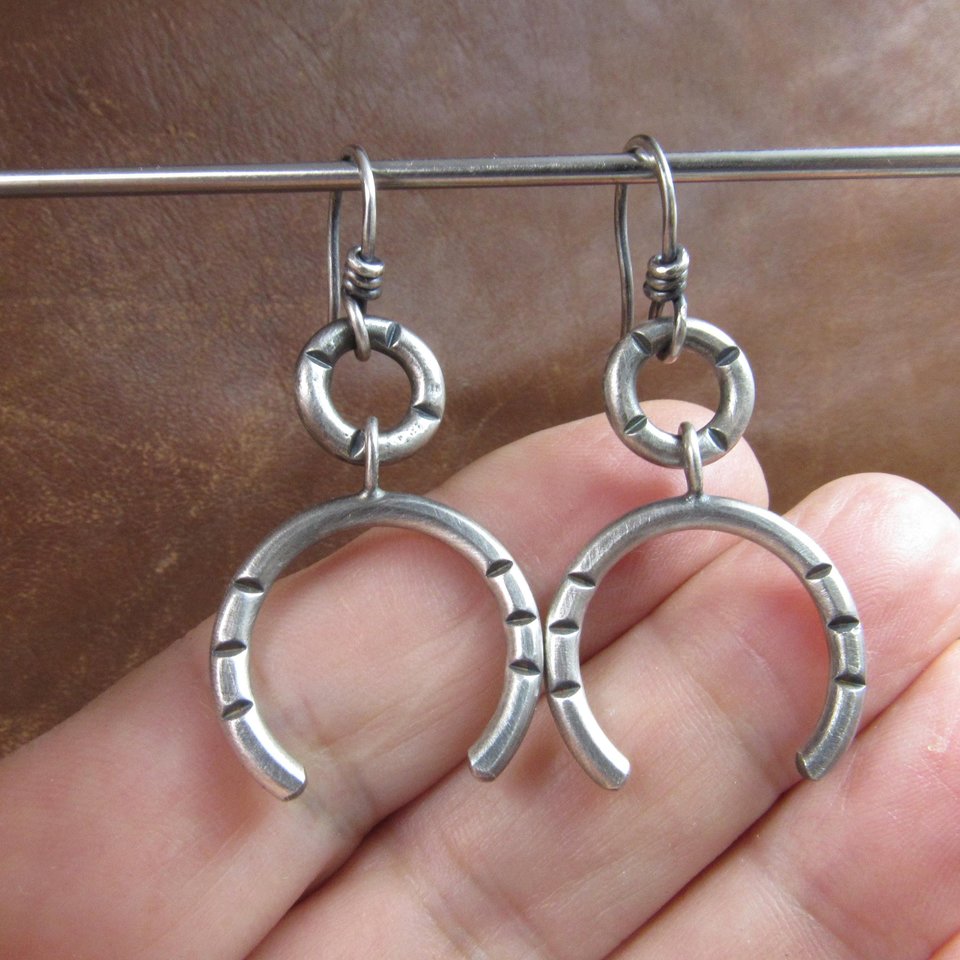 Close up of contemporary sterling silver earrings crafted from Argentium have lovely swingy movement. The double open hoop design allows for them to articulate. Lightweight and comfortable  at only 5 grams per earring. 1.75” long and 7/8” at widest point. Displayed against hand on brown leather.