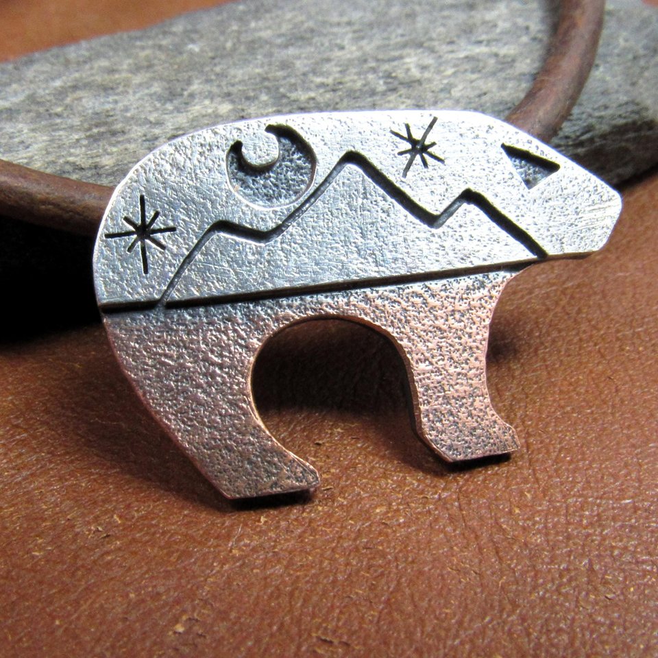 Bear pendant with the top half sculpted in silver with moon and star cutouts and the lower body in richly textured copper, joined harmoniously through traditional metalsmith techniques. The pendant is suspended on a sturdy 18-22 inch leather cord finished with our handmade Argentium silver findings for durability and comfort.Bear measures 1.25" long and is .75" tall. Displayed on brown leather against rock.