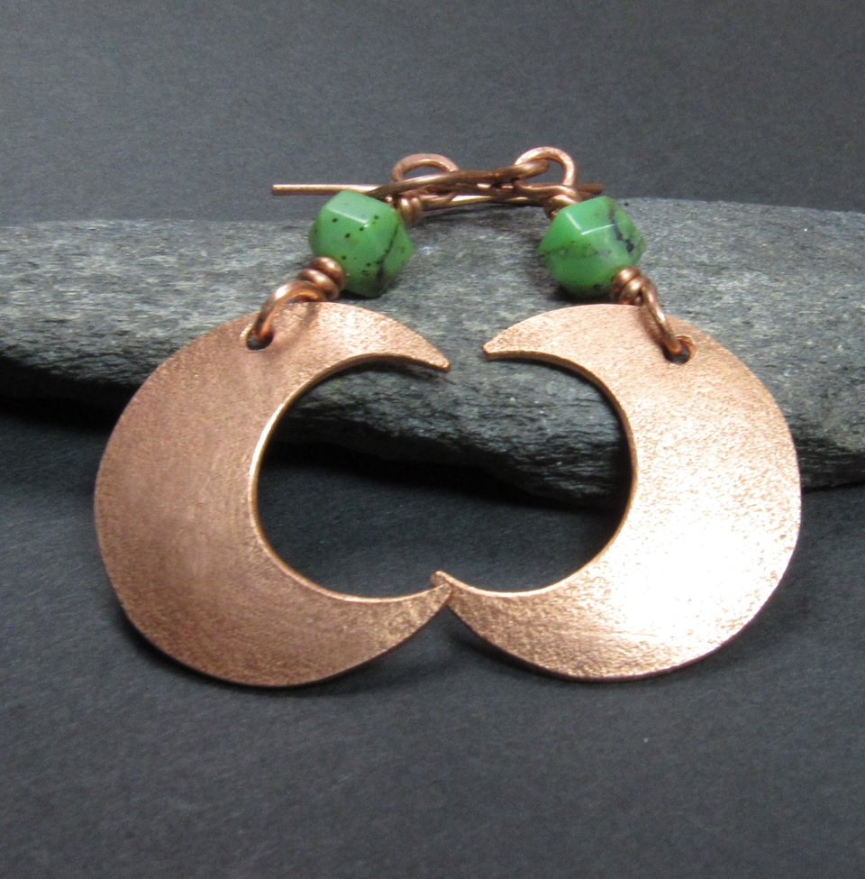 Up close photo of handmade copper crescent moon dangle earrings with faceted green chrysoprase beads. Earrings measure 2 and 1/8" long and the moon is 3/8" wide at widest point.The ear wires are solid, pure copper. Displayed against black rock on dark background.