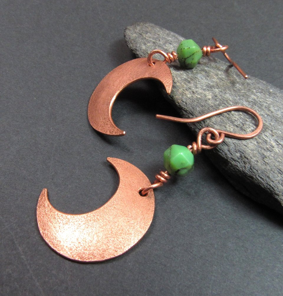 Side view of Handmade copper crescent moon dangle earrings with faceted green chrysoprase beads. Earrings measure 2 and 1/8" long and the moon is 3/8" wide at widest point.The ear wires are solid, pure copper. Displayed on dark background against rock.
