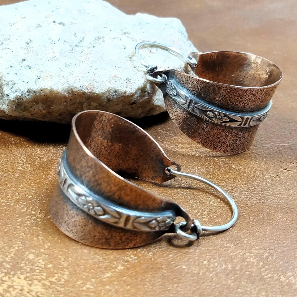 Copper and silver mixed metal basket shaped hoop earrings. The textured copper hoops are encircled with a floral pattern sterling silver wire and SS ear wires. Displayed on light brown back ground with white rock.