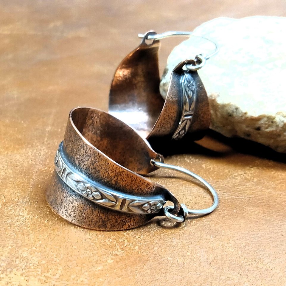 Close up of ear wire detail of Two tone mixed metal basket shaped hoop earrings in copper and sterling silver. The textured copper hoops are encircled with a floral pattern sterling silver wire and SS ear wires. Displayed on light brown back ground with white rock.