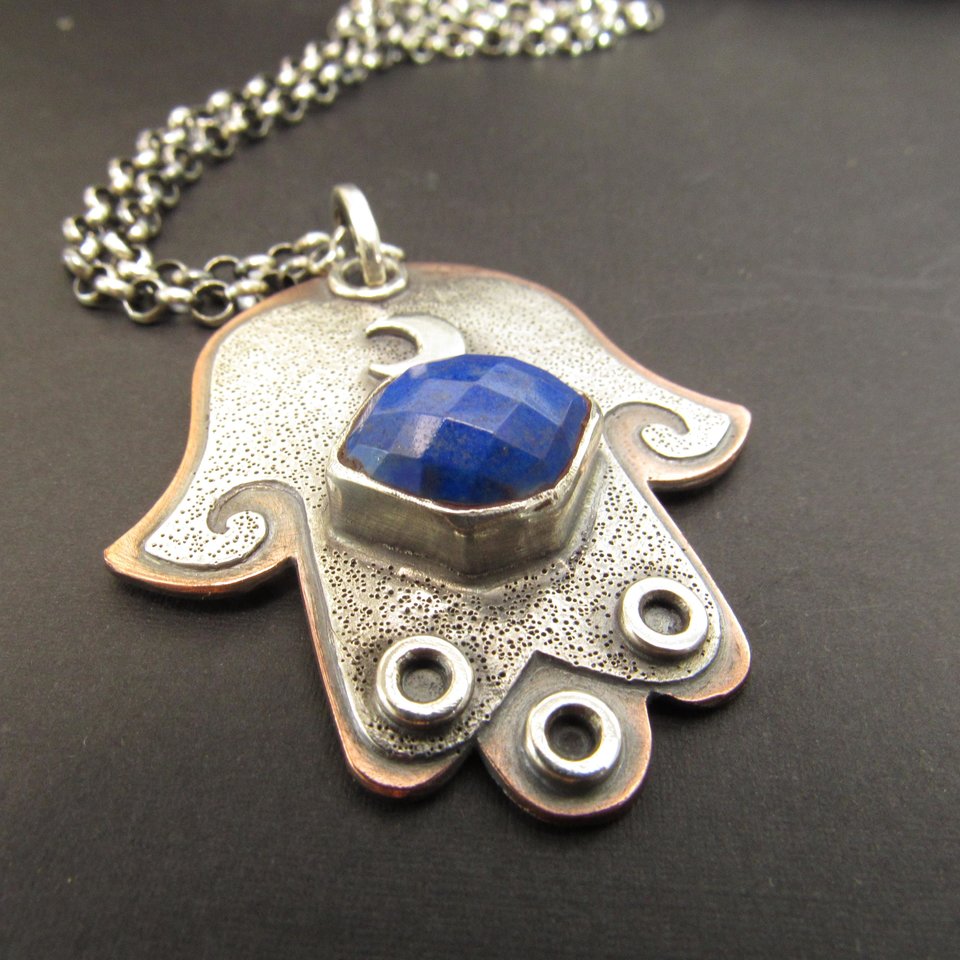 Mixed metal, copper and argentium sterling silver hamsa pendant necklace with lapis lazuli cabochon and crescent moon detail. 19 inch ss chain and findings. Hamsa is 1.5" wide x 1.75" tall. Displayed on dark grey background, pendant is facing forward.