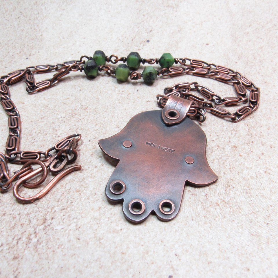 Reverse side of handcrafted copper, jade and chrysoprase hamsa pendant necklace showing makers mark "Mocahete". 2" tall x 1.5" wide, 24.5" copper scroll chain with 3 faceted chrysoprase beads on each side. All riveted design. Displayedon neutral background.