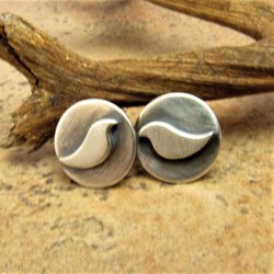 A pair of silver stud earrings with a bird design. The earrings are round and have a brushed and oxidized surface displayed against  a twig on sand colored tile.